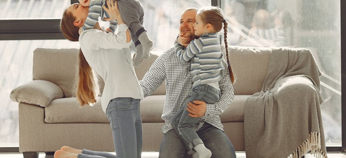Cheerful mother kissing little boy while father hugging adorable daughter when family sitting on floor in rainy day against window