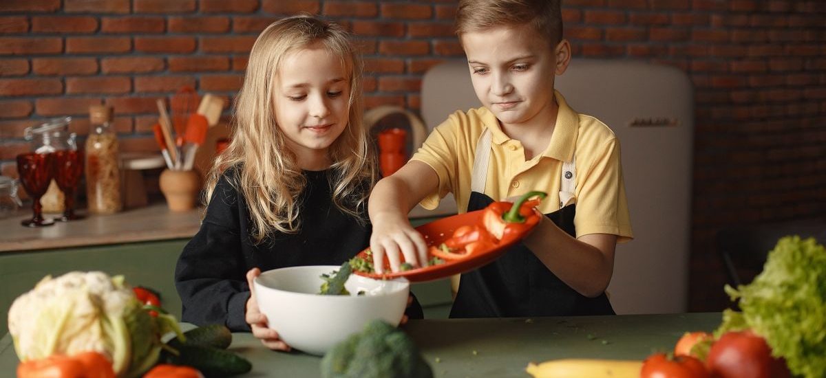 Little girl and boy mixing cutted pepper with other fresh ingredients for salad in big bowl while cooking at table with various fresh vegetables and fruits